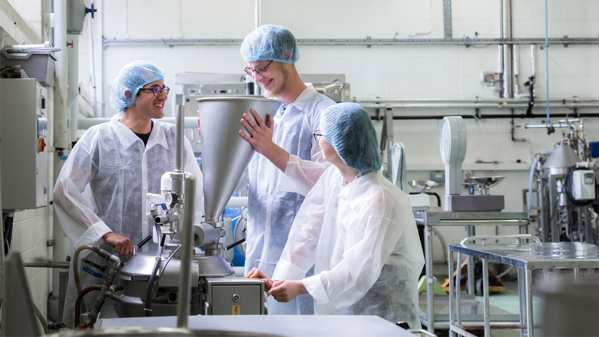 Three students in a food laboratory, wearing personal protective clothing. 